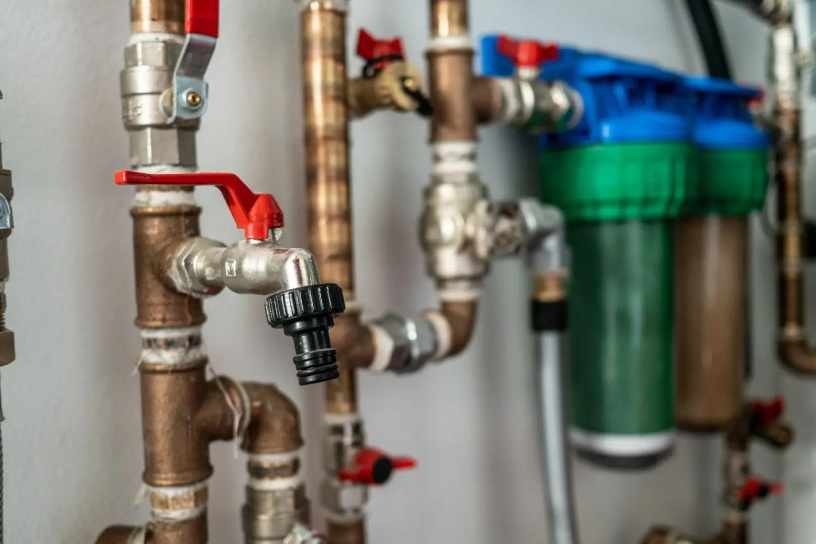 Close-up of interconnected copper pipes with red valve handles and a water filtration system mounted on a white wall.