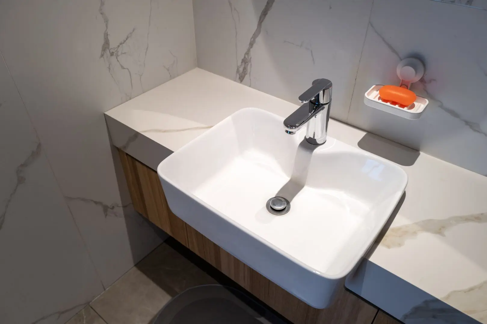 A modern white rectangular bathroom sink with a chrome faucet, set on a marble countertop. An orange bar of soap is in a wall-mounted holder above the sink.