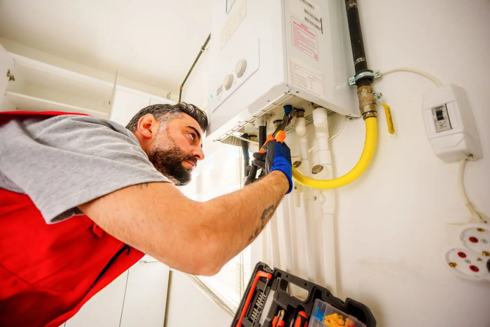 A technician in a red vest uses a screwdriver to repair or maintain a gas water heater unit mounted on a wall.