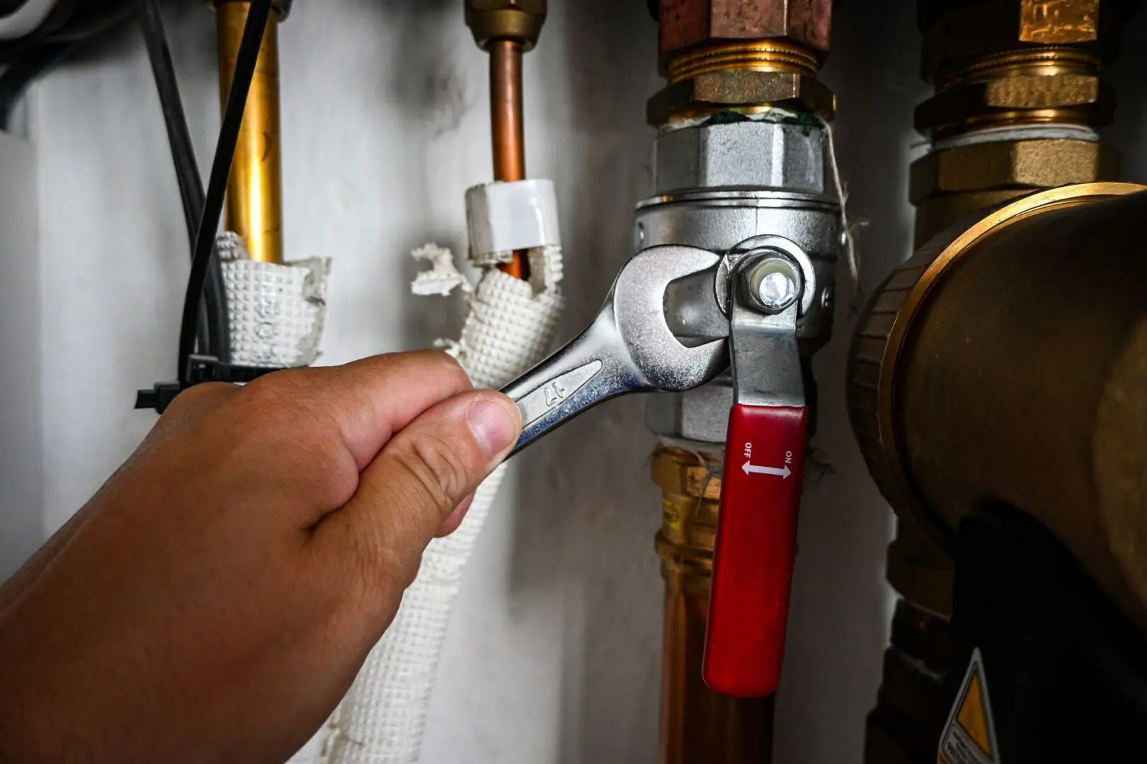 A hand uses a wrench to tighten a metal fitting on a pipe with a red-handled valve in a plumbing or heating system.