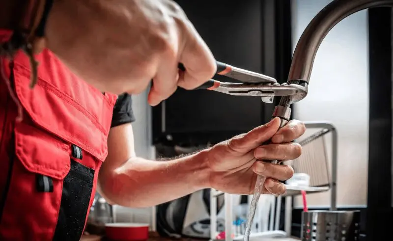 Person using pliers to tighten or adjust a kitchen faucet while water flows from the tap.
