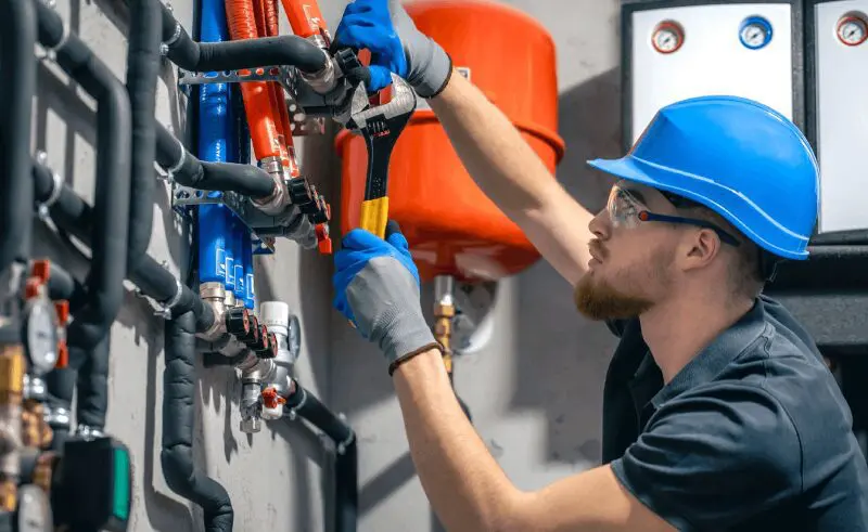 A worker in a blue hard hat adjusts pipes with a wrench in a utility room, surrounded by valves and equipment.