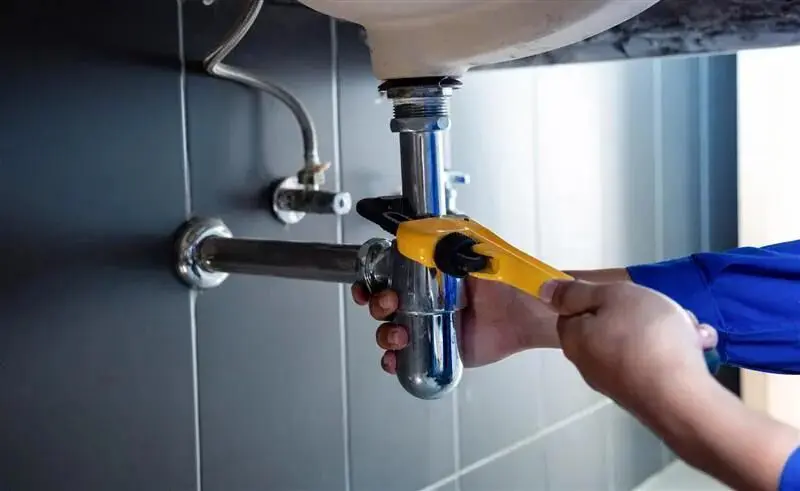 A person uses a wrench to tighten or loosen a pipe under a sink, performing plumbing maintenance.