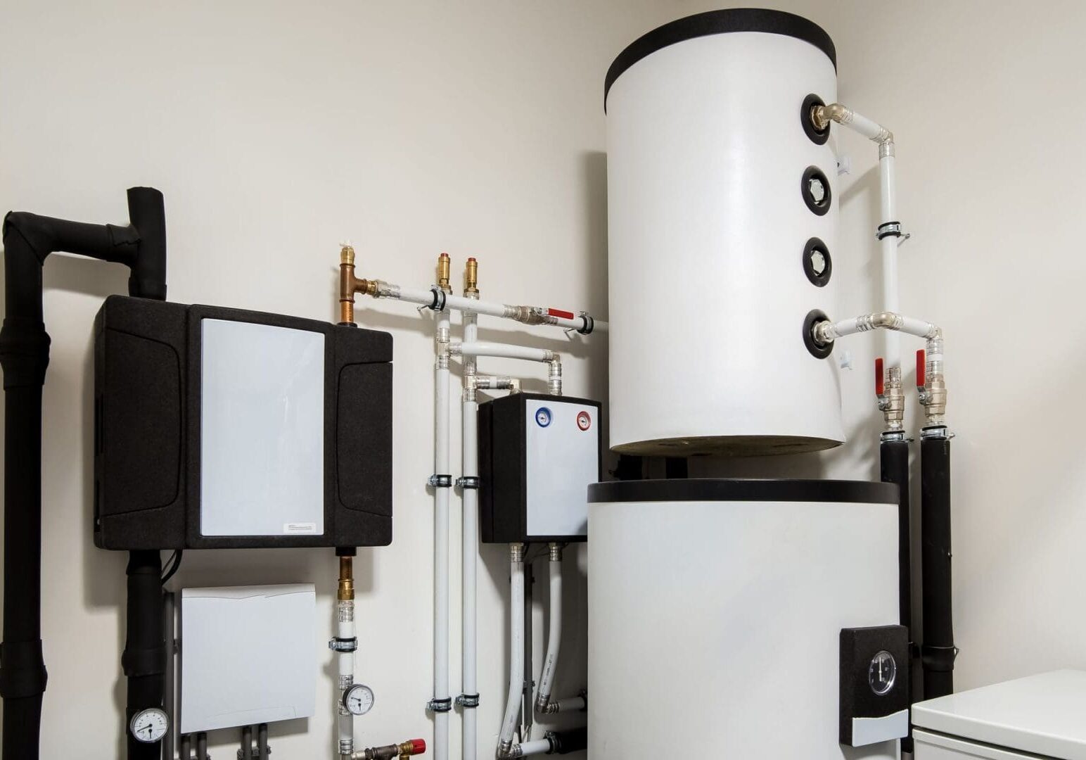 Modern residential heating system with a large cylindrical water tank, insulated pipes, pressure gauges, and control panels mounted on a wall.