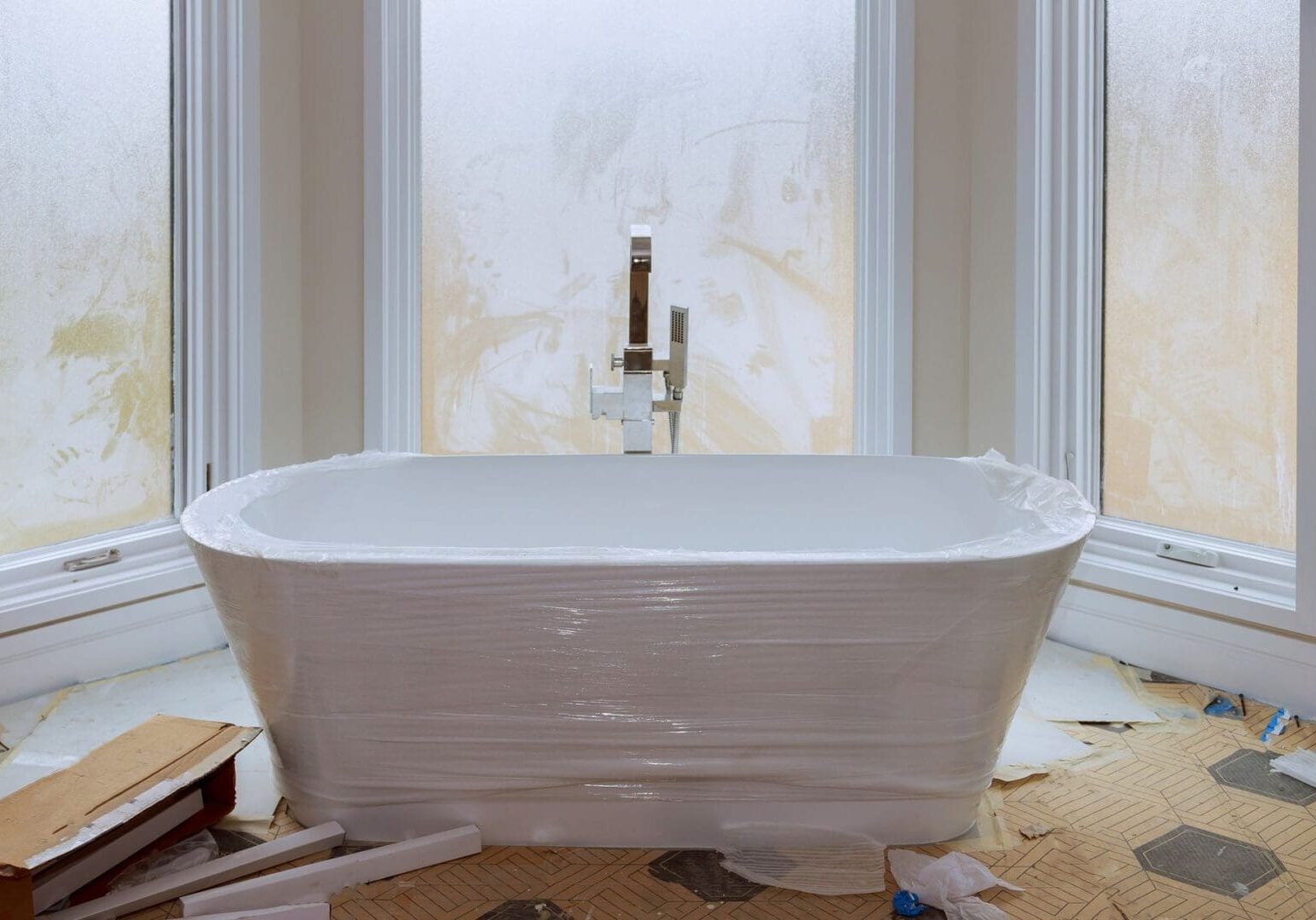 A white bathtub wrapped in protective plastic sits in front of three frosted windows; the surrounding floor and area appear to be under renovation.