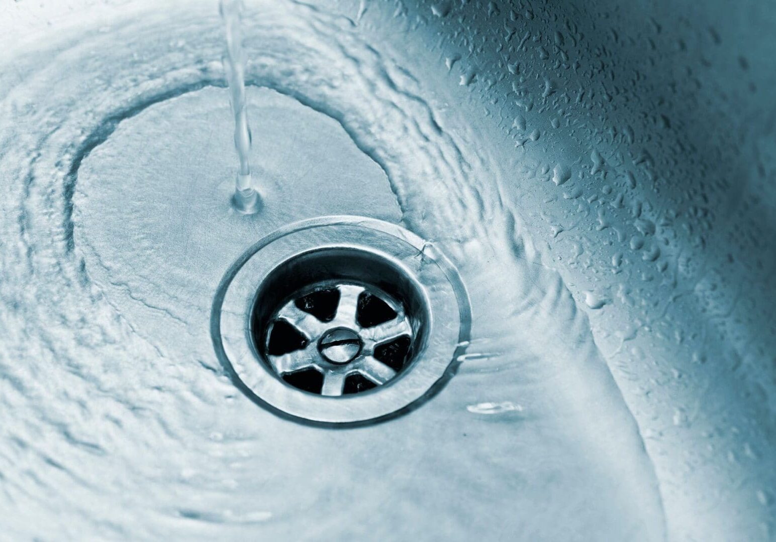 Close-up of water flowing into a metal sink drain, with water droplets and ripples visible around the drain area.