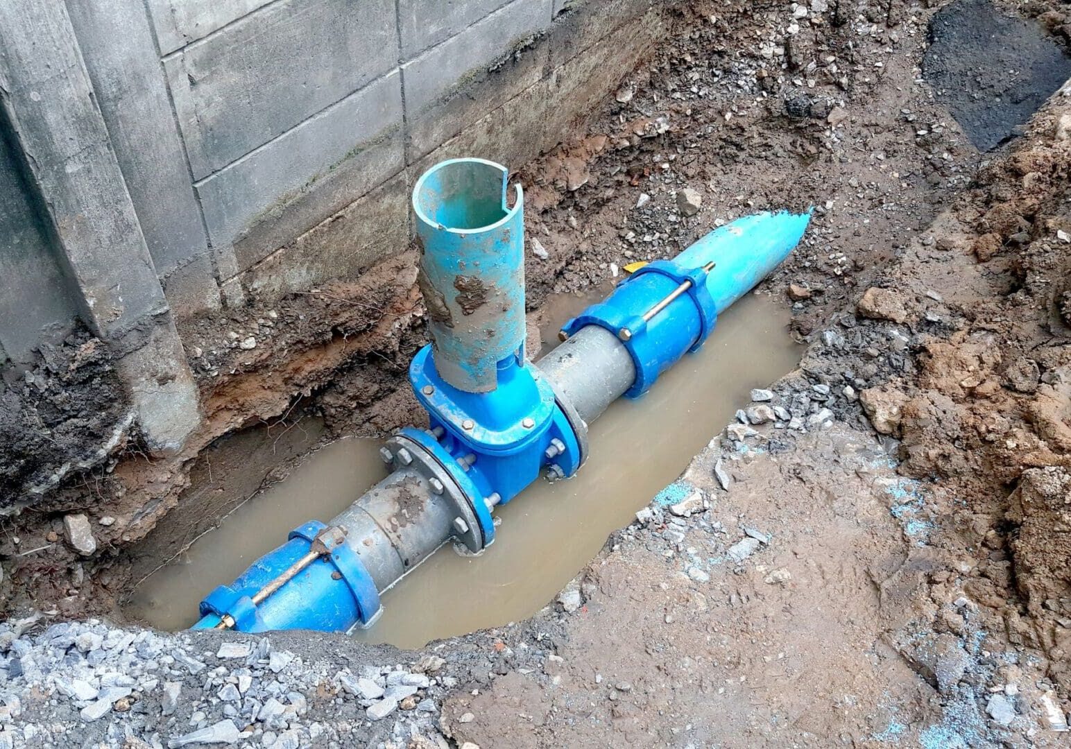 Exposed underground blue water pipe with a vertical connector in a muddy trench next to a concrete wall.