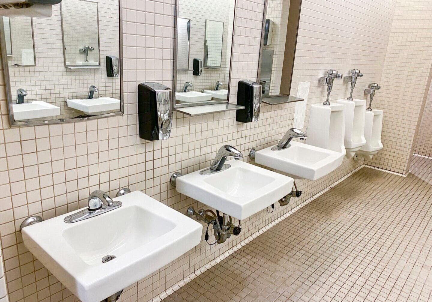 A public restroom with three sinks, mirrors, soap dispensers, and three urinals along a tiled wall and floor.