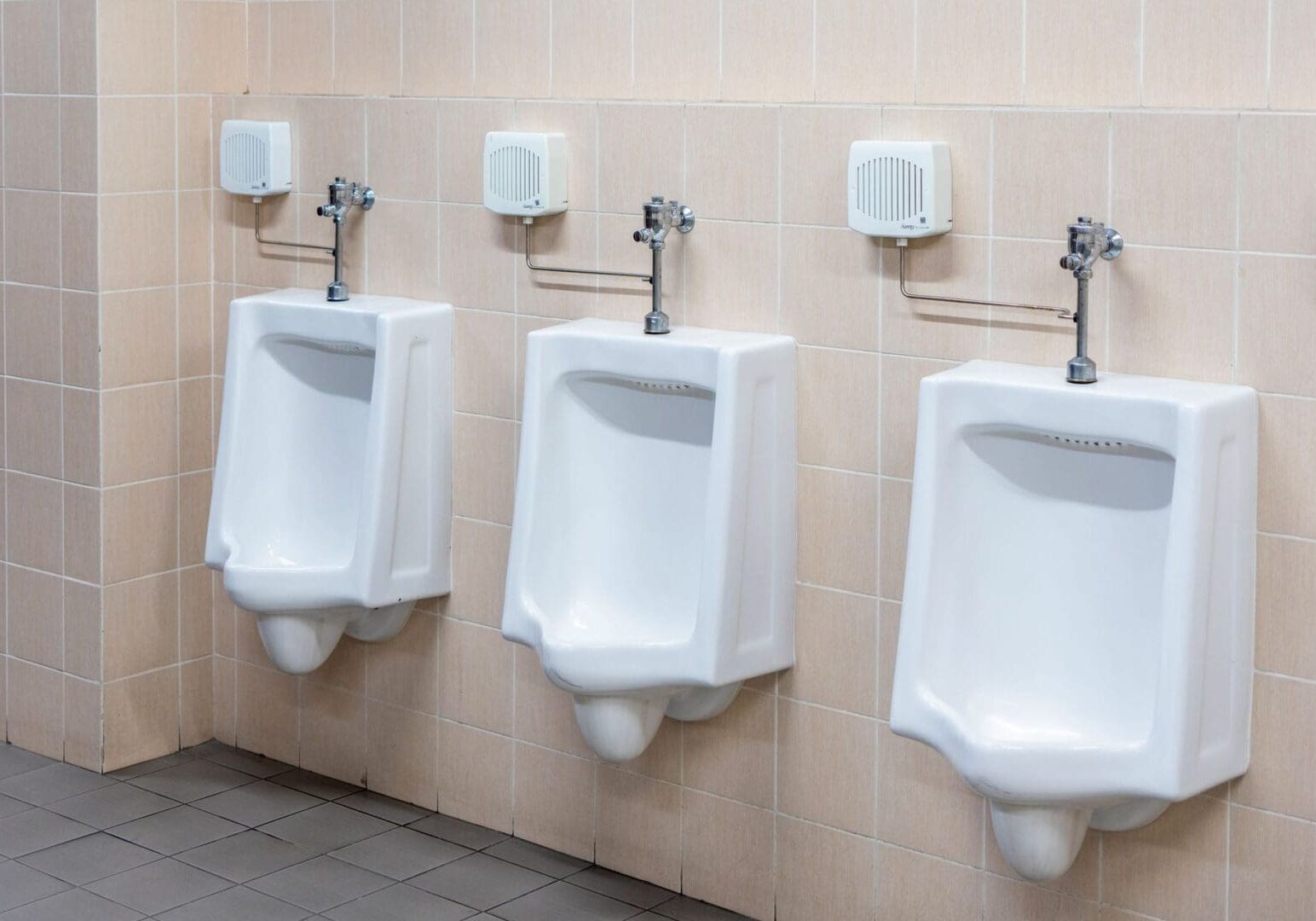 Three white ceramic urinals are mounted on a beige tiled wall in a public restroom with gray tiled flooring.