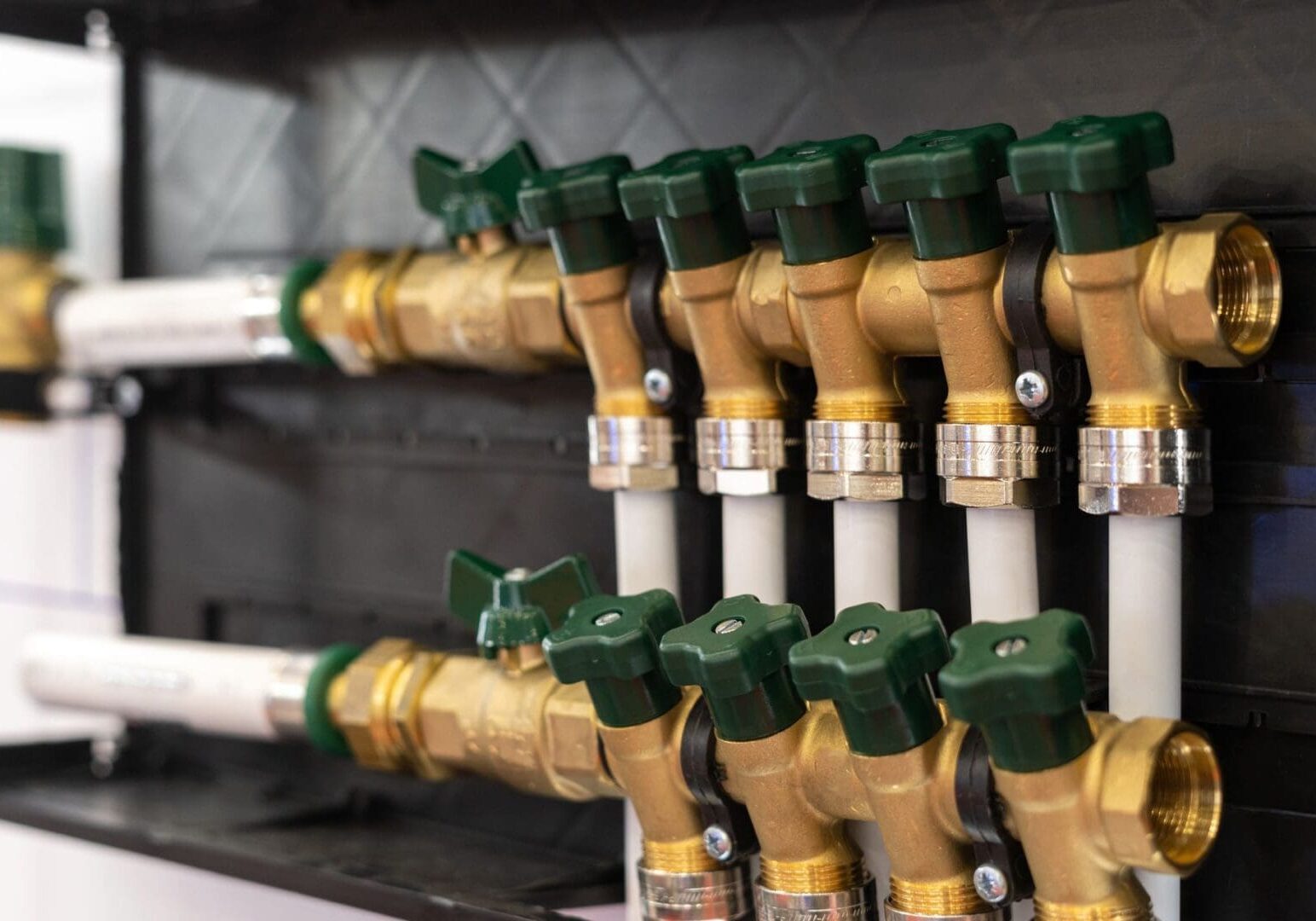 Close-up of a row of brass water valves with green handles connected to white plastic pipes, mounted on a black panel.