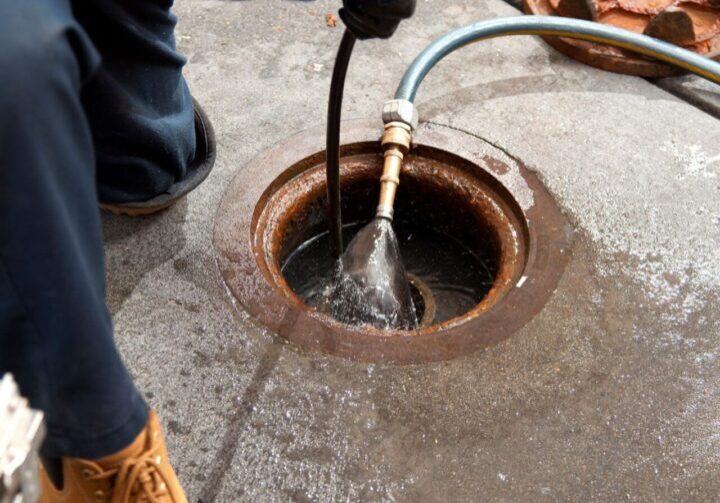 Person in gloves and work boots using a hose to clean or unclog an industrial drain on a concrete floor.