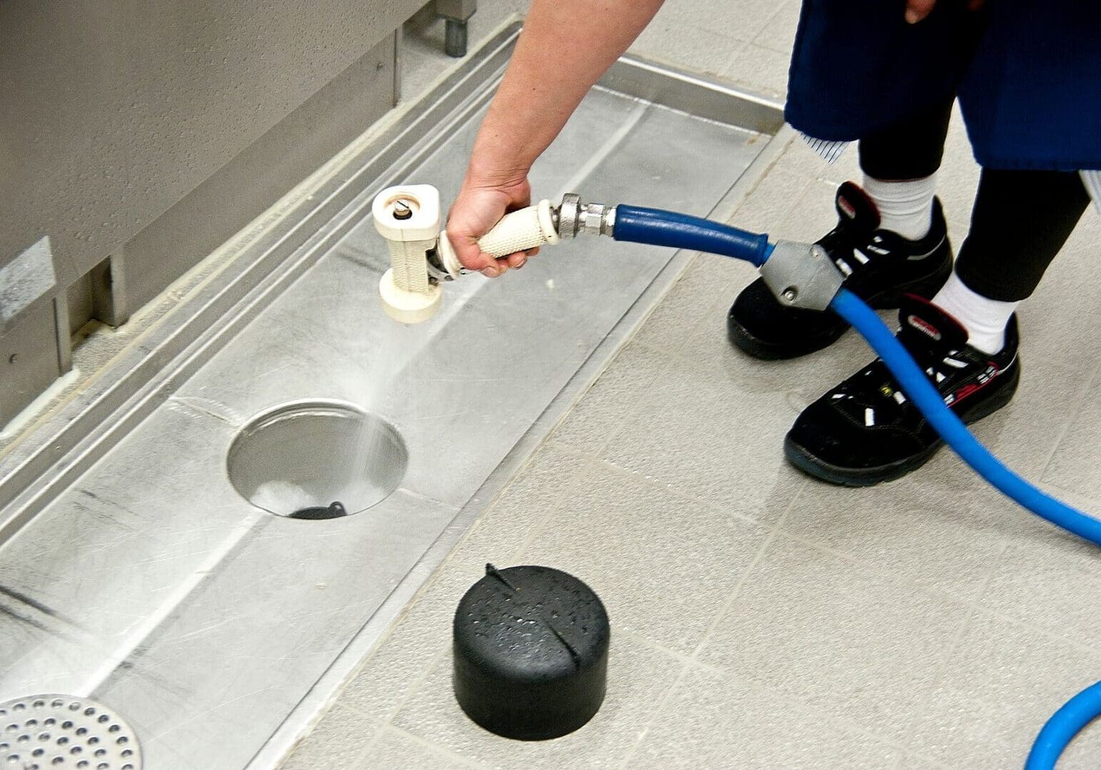 A person uses a hose with a spray nozzle to clean a floor drain in a tiled setting. A black cap is placed nearby on the floor.