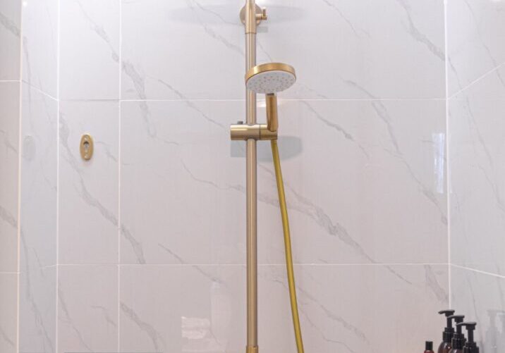 A modern shower setup with gold fixtures, a rainfall showerhead, a handheld showerhead, and a yellow hose on a white marble tiled wall. Black soap dispensers and toiletries are on a wall shelf.