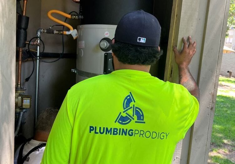 Two workers, one wearing a "Plumbing Prodigy" shirt, install or service equipment in a small outdoor utility area with pipes and a water heater.