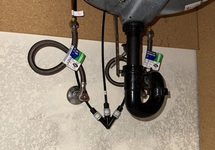 Pipes and hoses with labels are visible under a kitchen sink, attached to plumbing and drainage fixtures against a white wall.